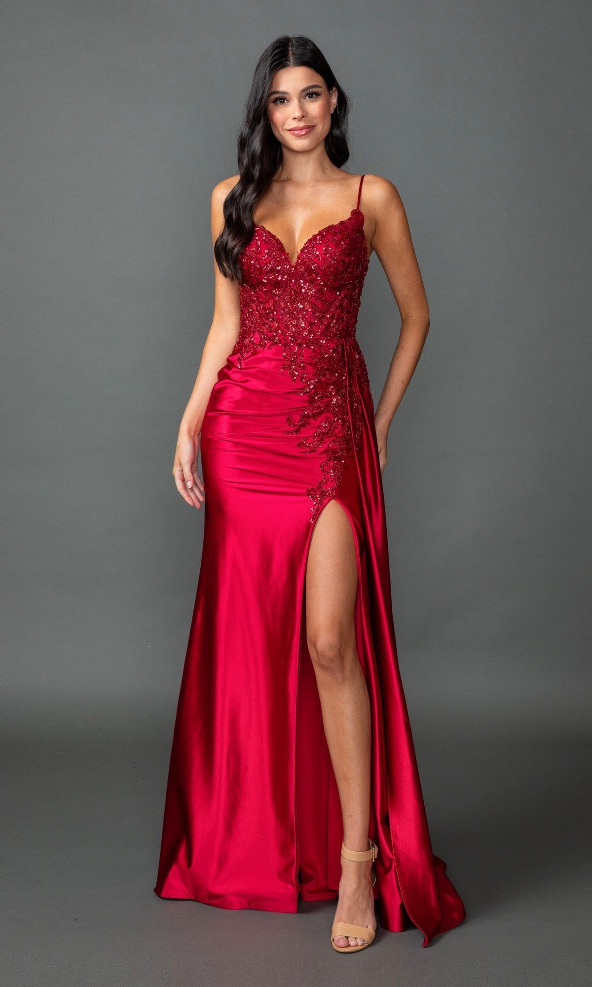A woman wears the Dancing Queen 4552 long prom dress in burgundy, featuring beaded embroidery, lace details, spaghetti straps, and a thigh-high slit, standing against a plain grey background.